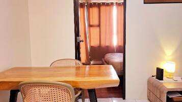 Una stanza con un tavolo da pranzo in legno, due sedie in vimini, un piccolo comodino con una lampada e una porta che dà su una camera da letto con un letto e tende.