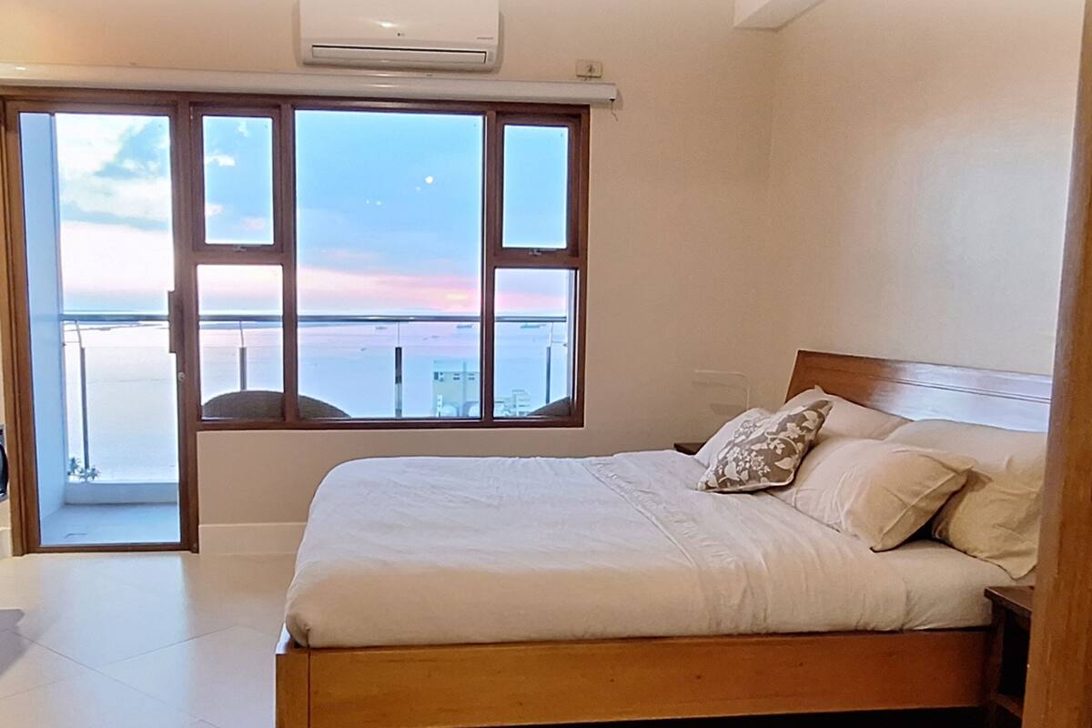 A bedroom with a wooden bed, white bedding, and a view through large windows.