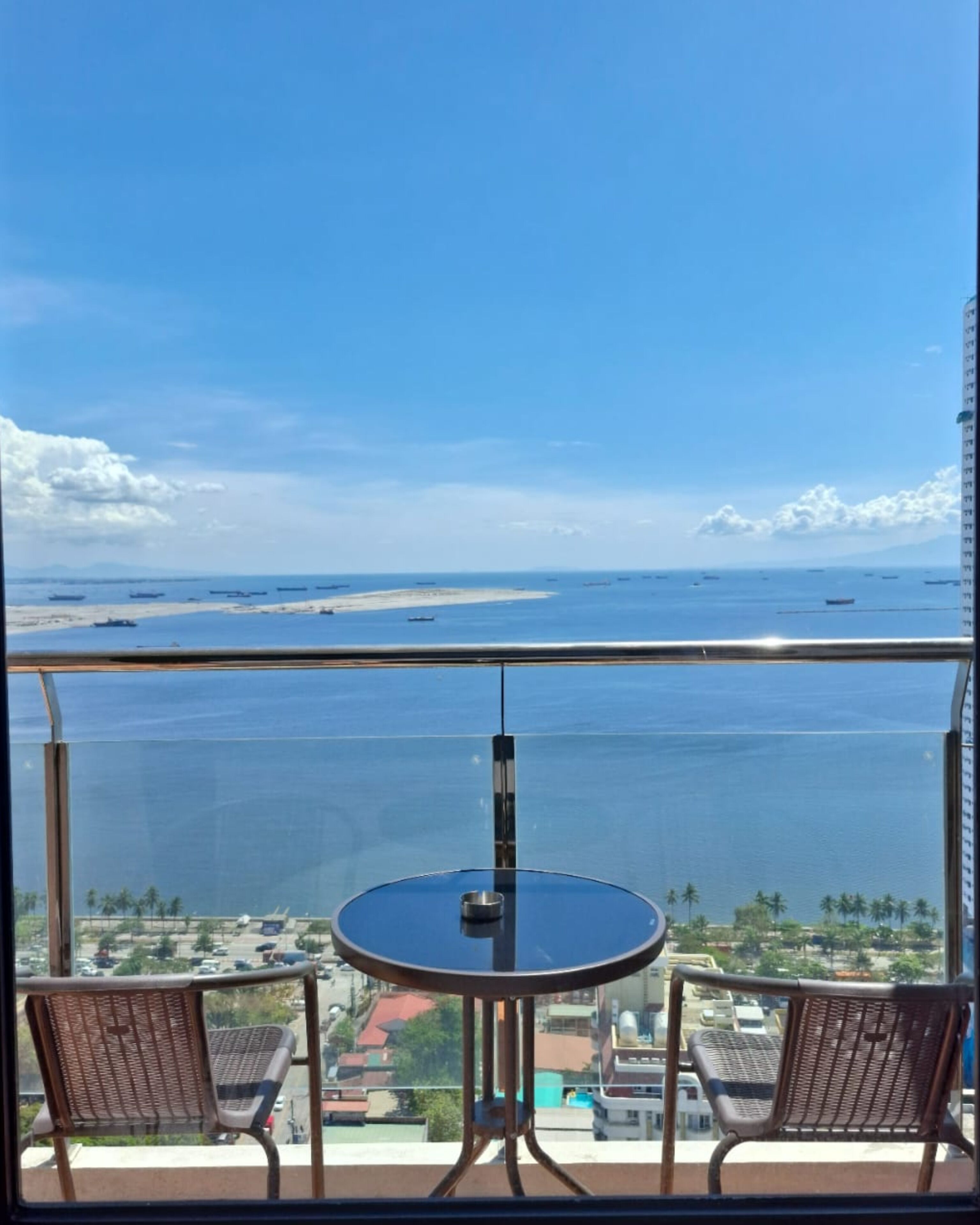 Ein Balkon mit Tisch und zwei Stühlen, mit Blick auf ein Gewässer, in dem Schiffe fahren, und eine entfernte Küstenlinie.