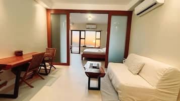 A compact living space with a sofa, a dining table with chairs, a small coffee table, and a bed in the background.
