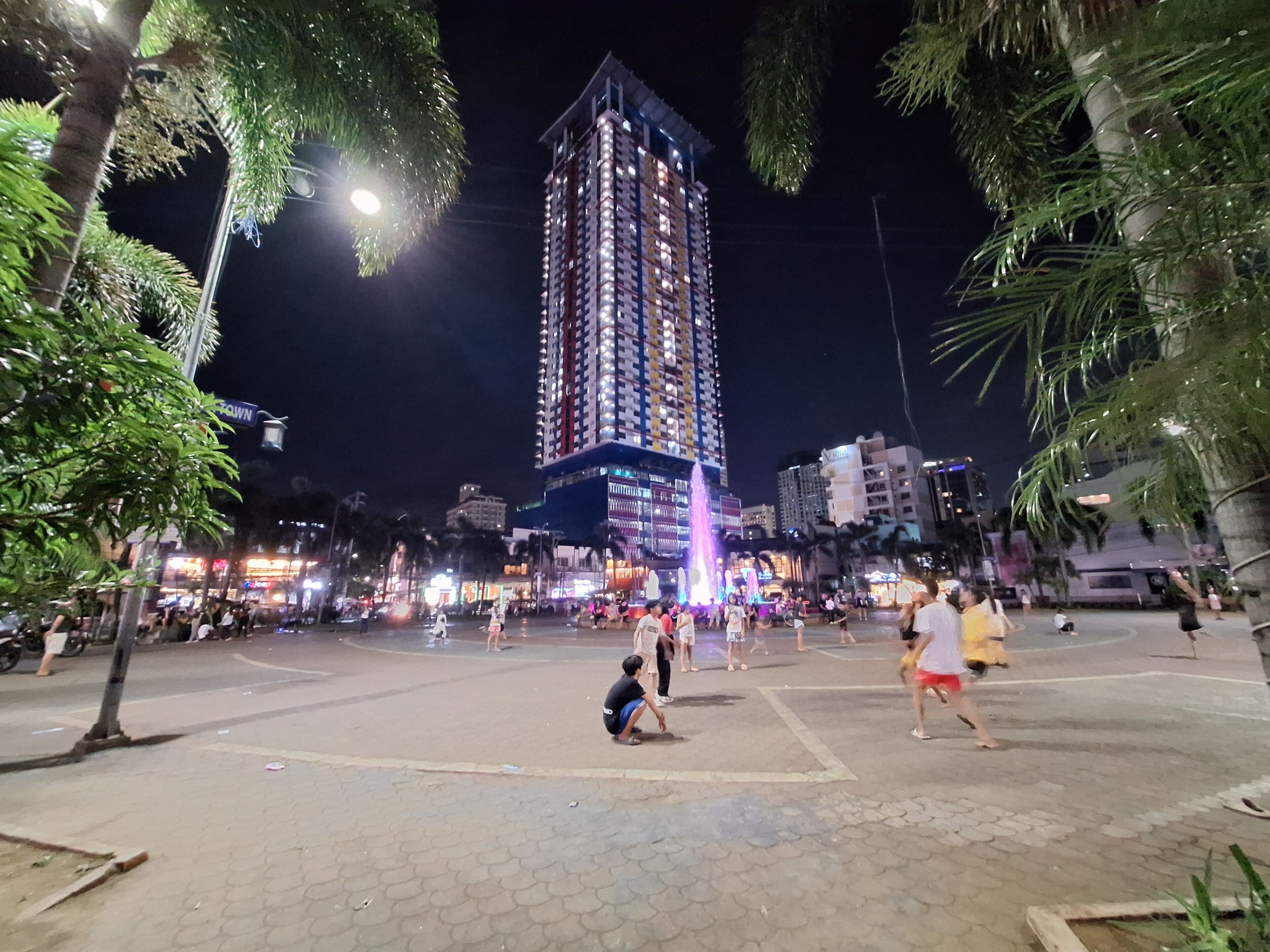 A nighttime urban scene with a tall illuminated building, a fountain, and people in a public square.