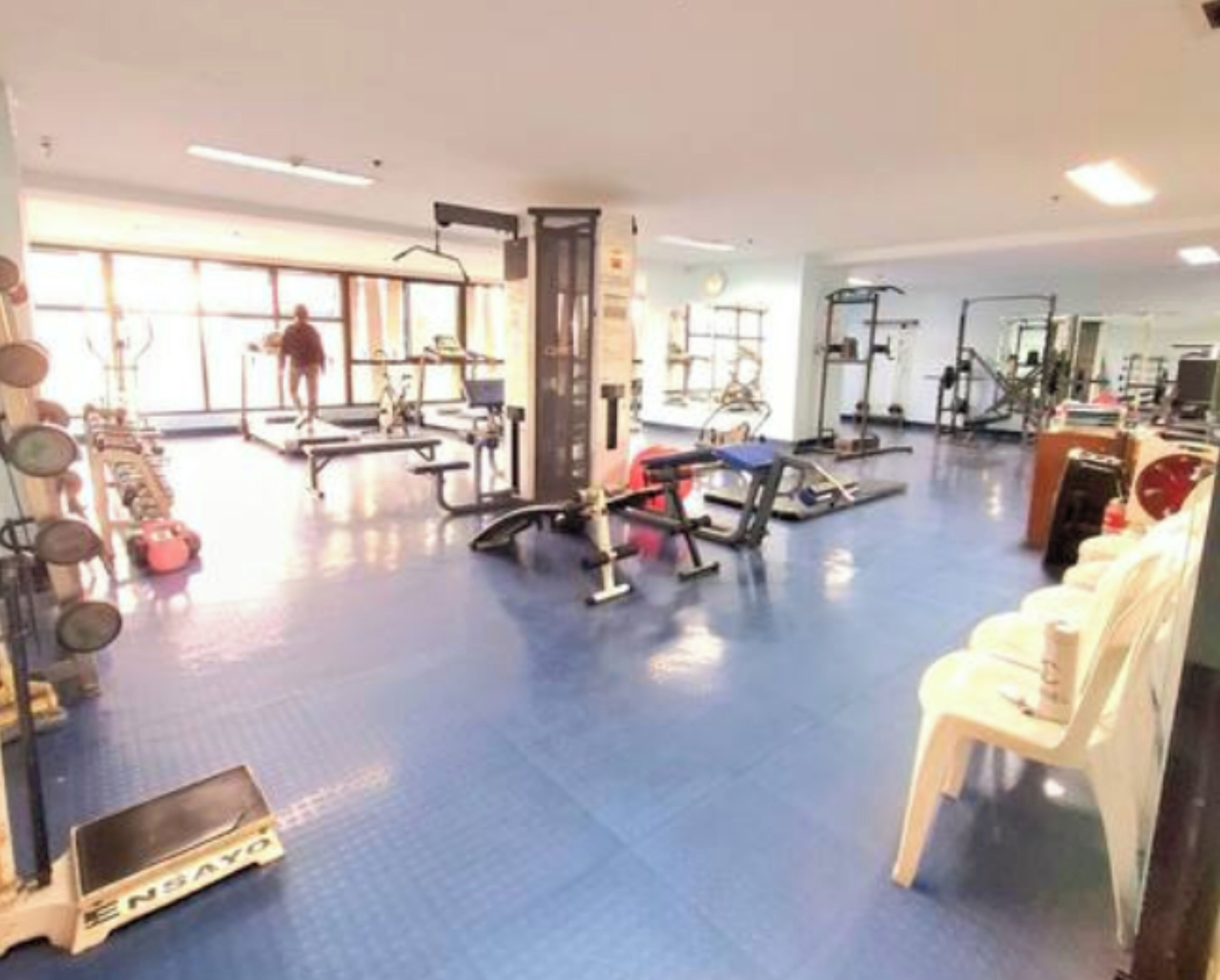 Une salle de sport équipée de divers appareils d’exercice, avec un sol bleu, et une personne qui s’entraîne.