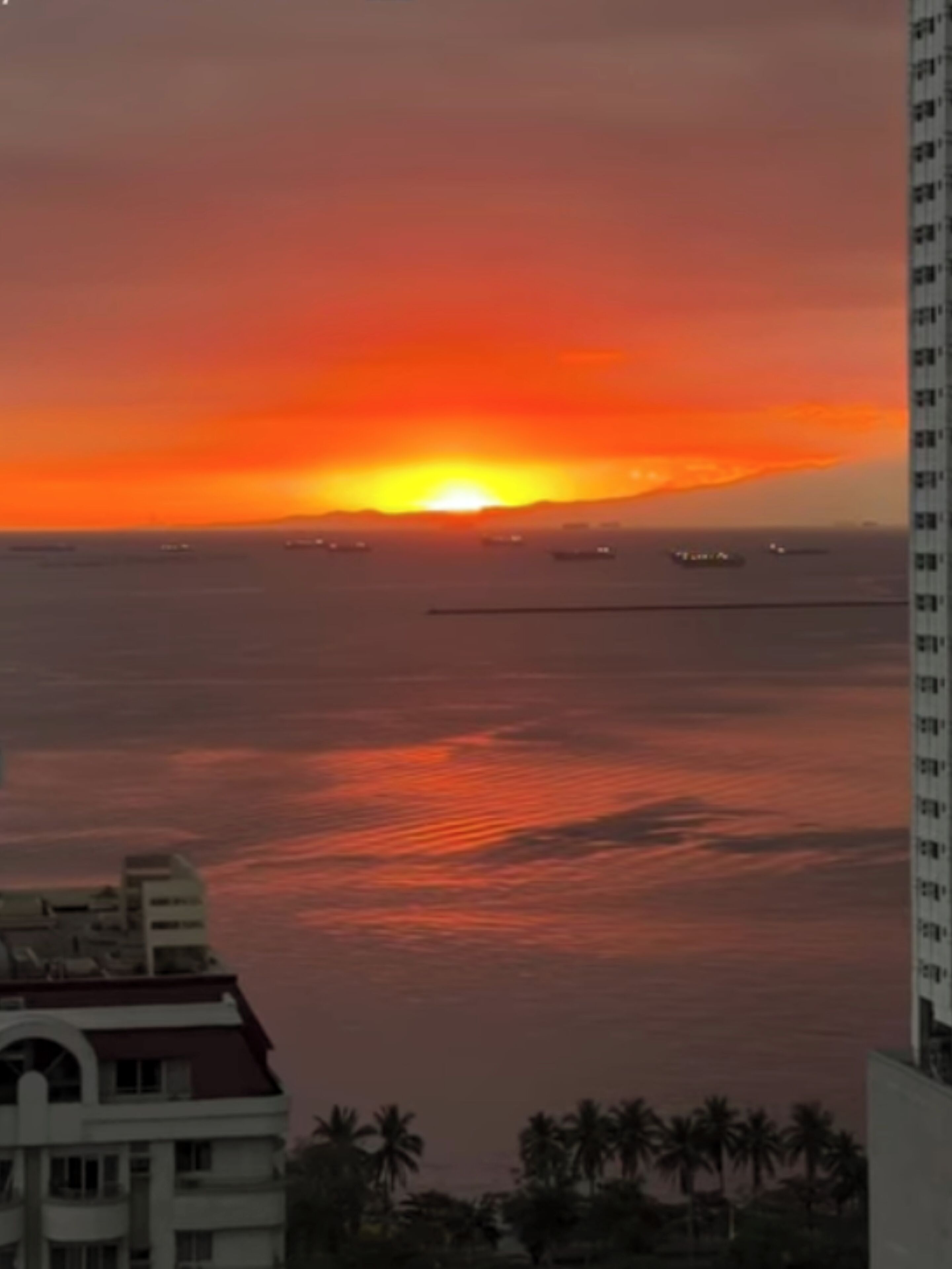 A sunset over a calm sea with a distant view of a city skyline and a tall building on the right.