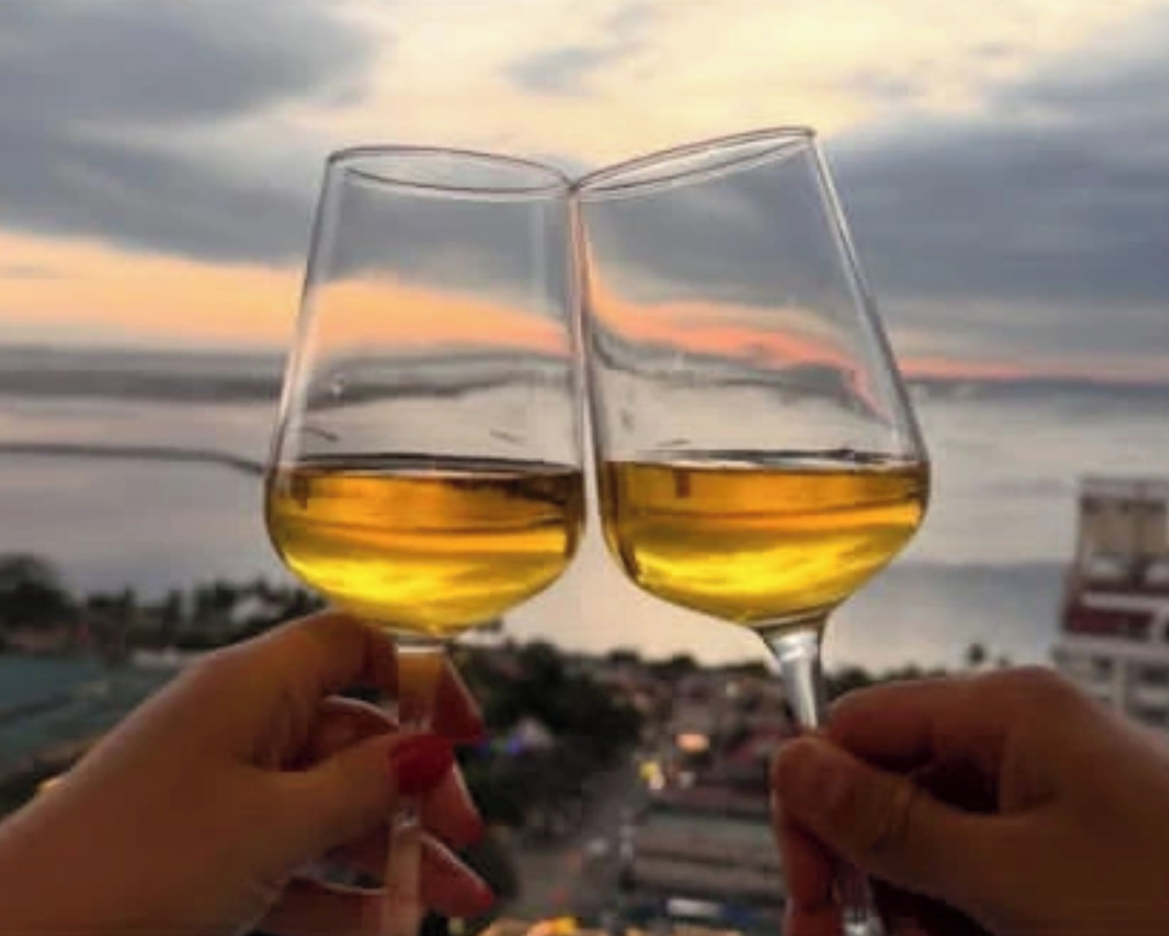 Dos manos sosteniendo copas de vino con una vista al atardecer de fondo.