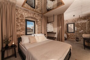 A bedroom with a stone wall, a bed with a textured blanket, a bedside table with a lamp, and a window with a view of buildings. - Villa Imagination with Pool (Gornji Humac)