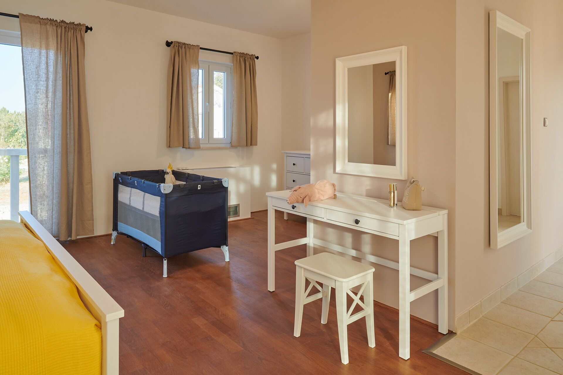 A bedroom with a bed, a small white desk with a mirror, a bedside table, a window with curtains, and a balcony.