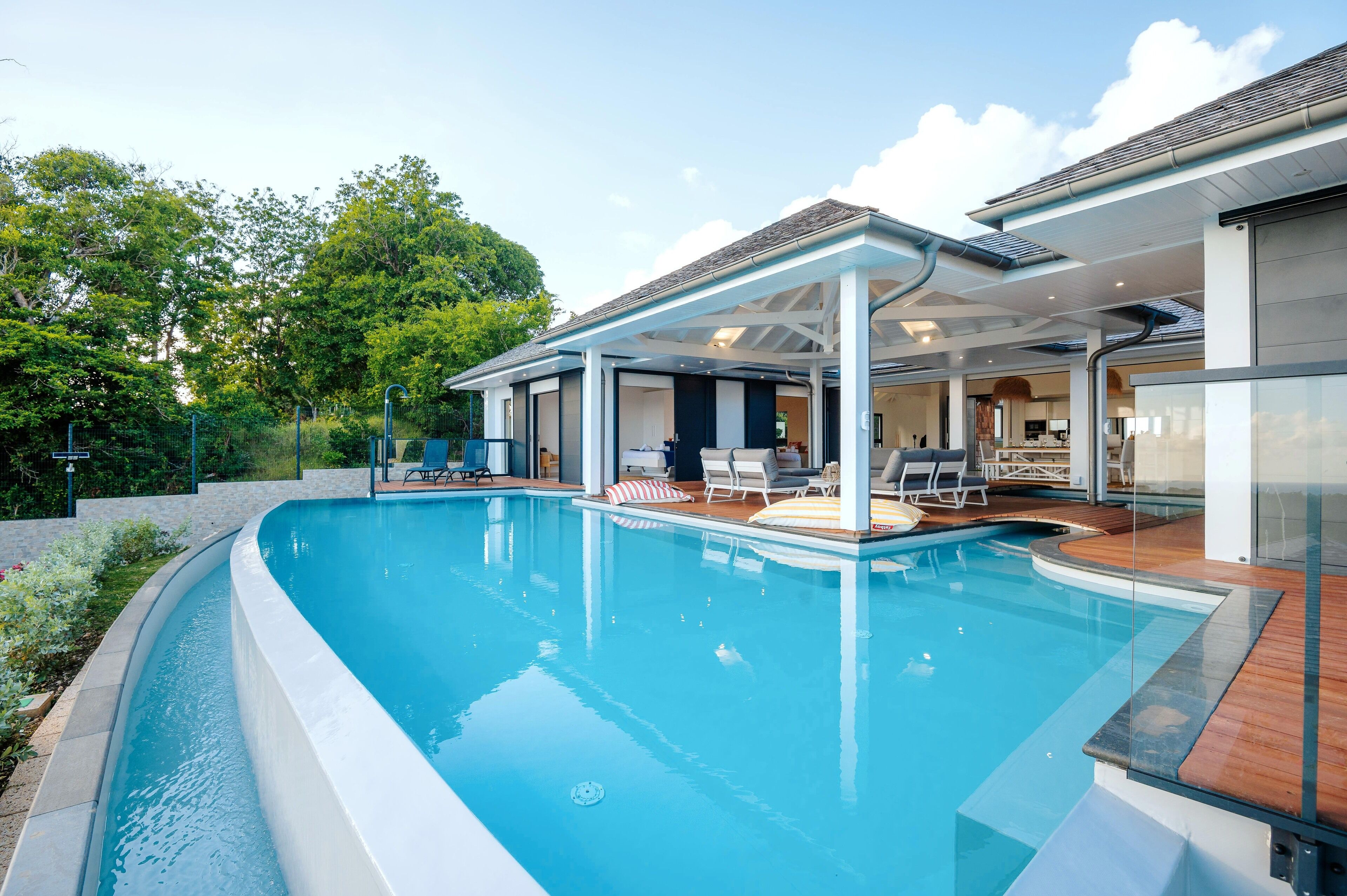 En moderne udendørs swimmingpool med en overdækket terrasse, liggestole og udsigt til huset.