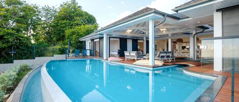 En moderne udendørs swimmingpool med en overdækket terrasse, liggestole og udsigt til huset.