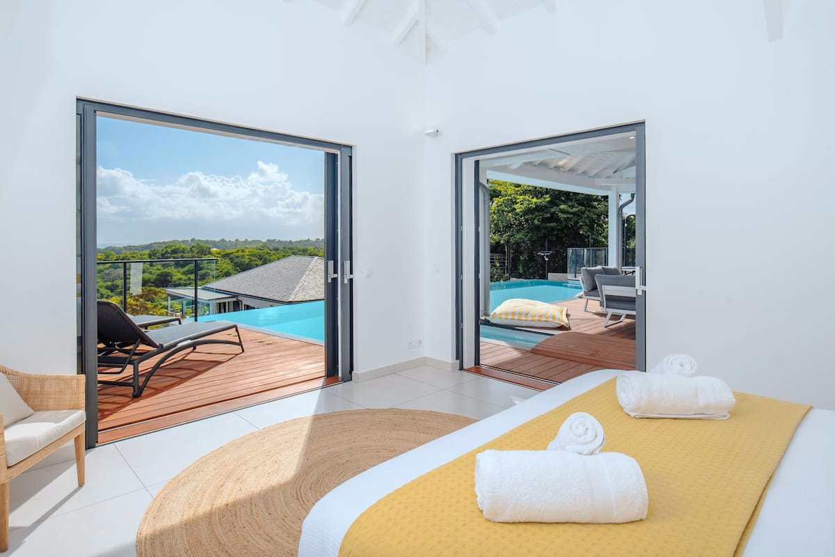 A modern hotel room with a large bed, a wooden deck, and a view of a swimming pool and greenery.