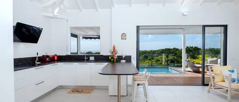 Cocina moderna con una isla central, televisor de pantalla plana y comedor con vistas a una piscina y vegetación.