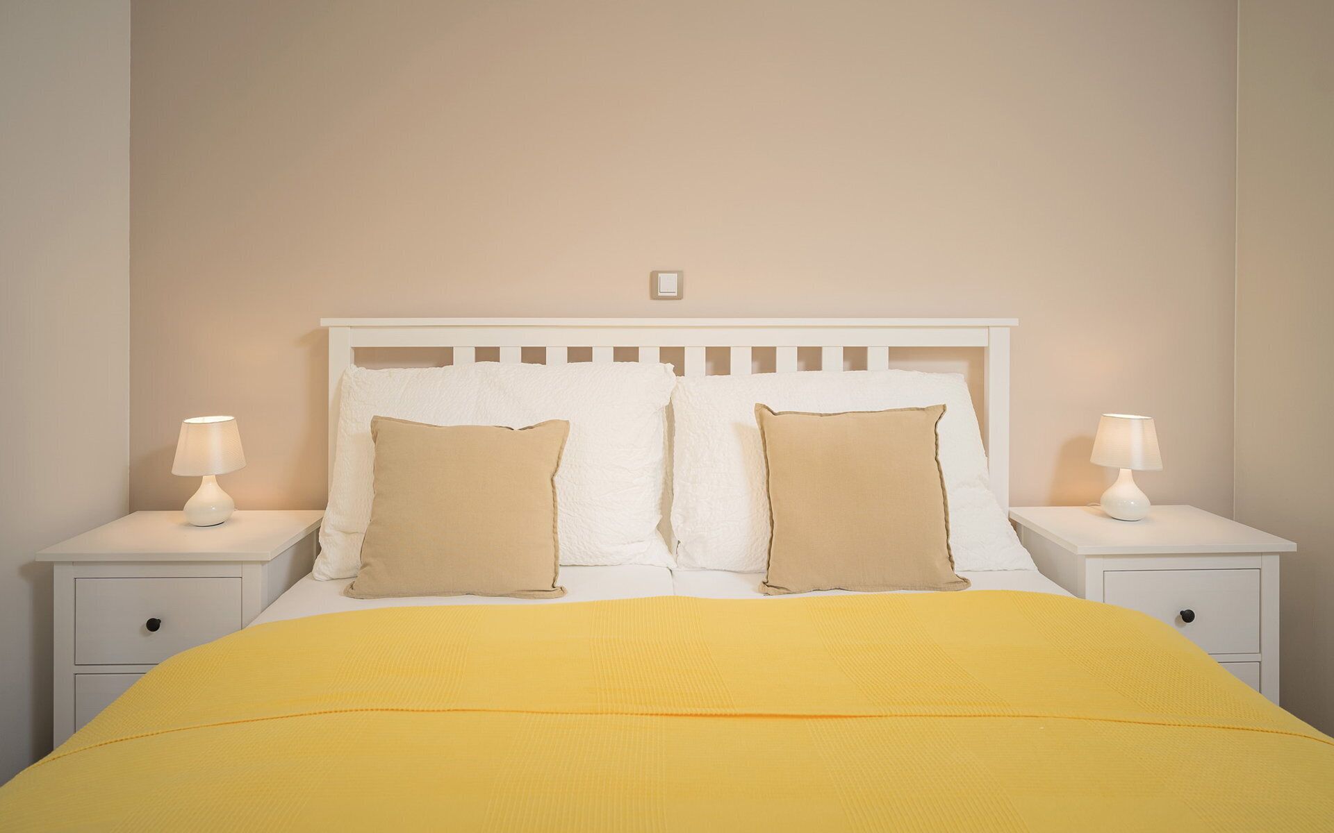 Een bed met een wit hoofdbord, twee nachtkastjes met lampen en een gele sprei.