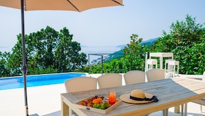 A wooden table with wicker chairs, a tray of fruit and pastries, and a drink, set against a backdrop of a swimming pool and lush greenery. - Villa Piano with Pool (Matulji)