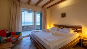 A bedroom with a large bed, a wooden headboard, a nightstand, and a view of the sea through a sliding glass door.