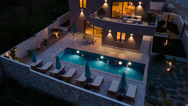 A modern two-story house with a swimming pool, outdoor seating, and a well-lit interior.
