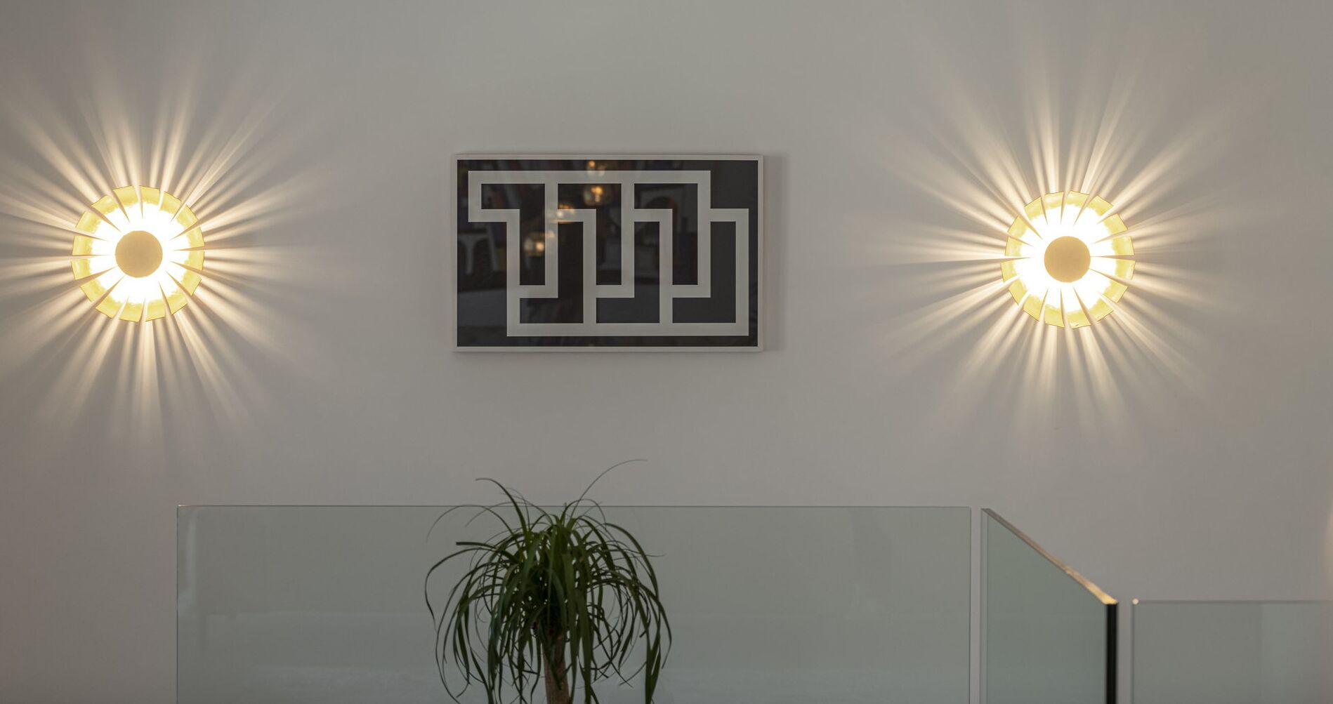 A reception area with two wall-mounted lights, a geometric wall art piece, and a potted plant.