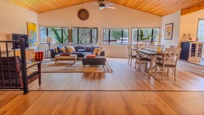 A spacious living room with a high ceiling, wooden flooring, a sofa set, a dining table with chairs, and large windows offering a view of the outdoors.