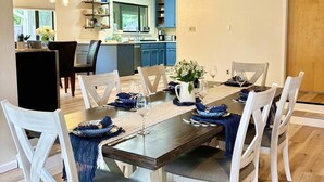 A dining area with a wooden table set for six, white chairs, and a kitchen in the background.