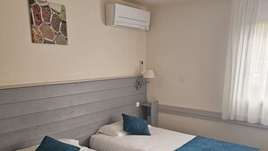 Habitación de hotel con dos camas, aire acondicionado de pared, lámpara de noche y ventana con cortinas translúcidas.