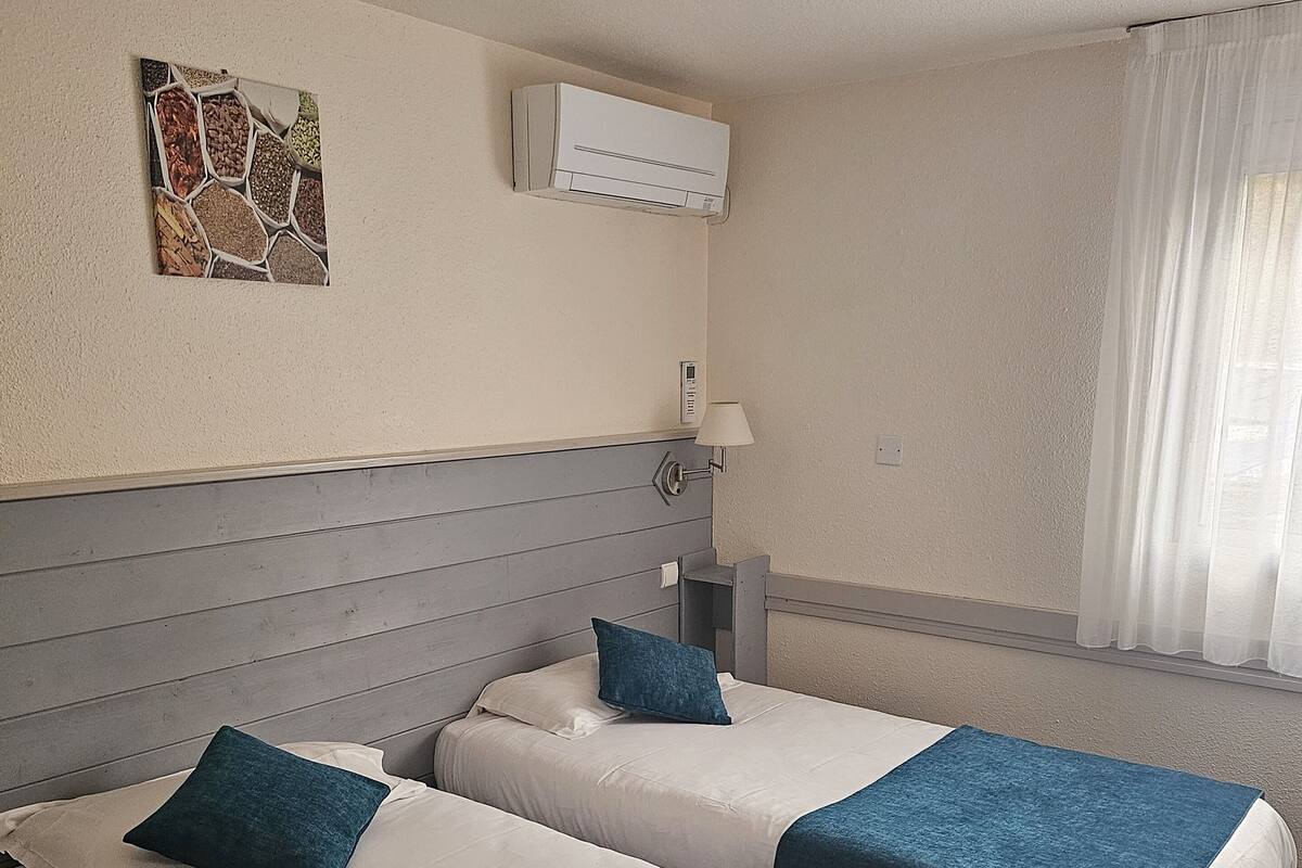 Une chambre d’hôtel avec deux lits, un climatiseur fixé au mur, une lampe de chevet et une fenêtre avec des rideaux légers.