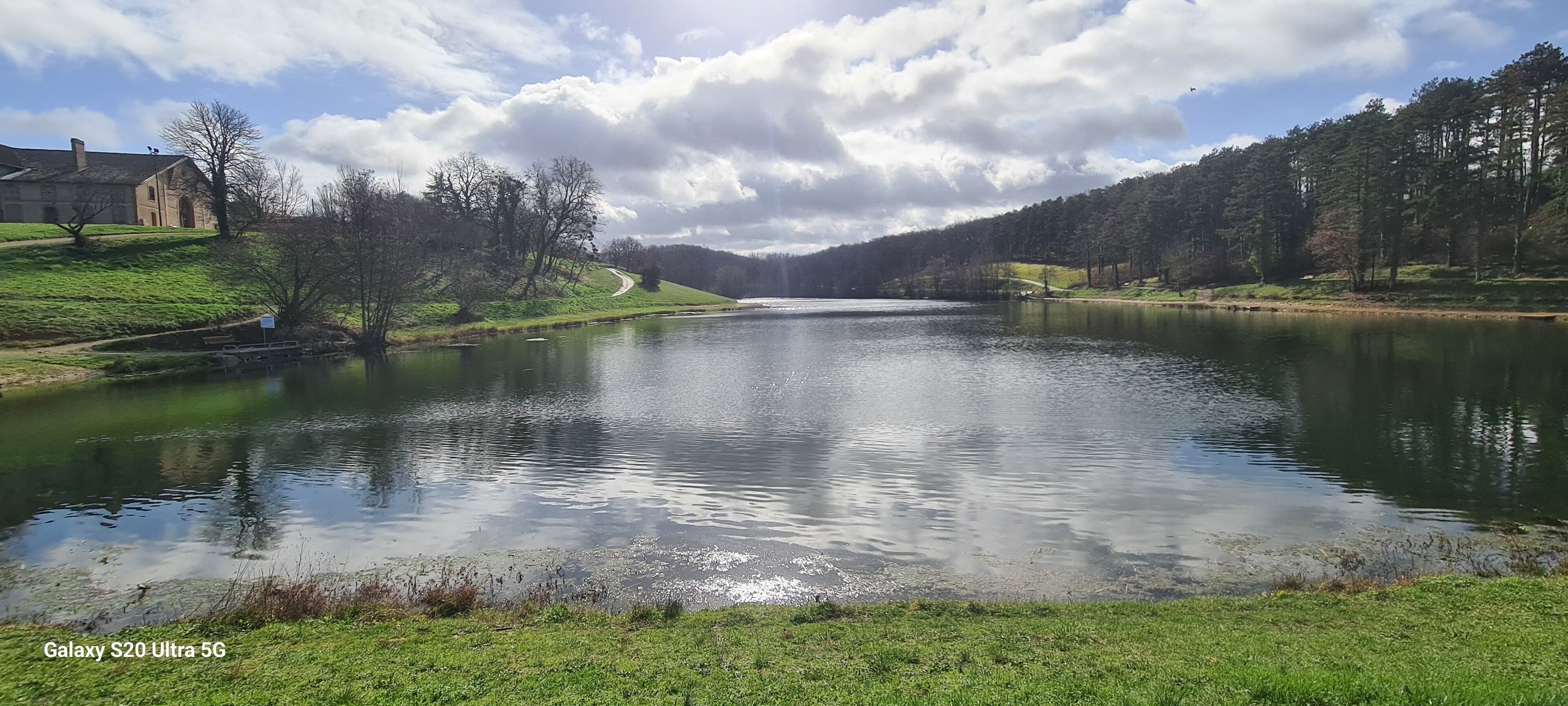 Um lago sereno com uma margem gramada, um edifício de pedra à esquerda e uma colina arborizada à direita.