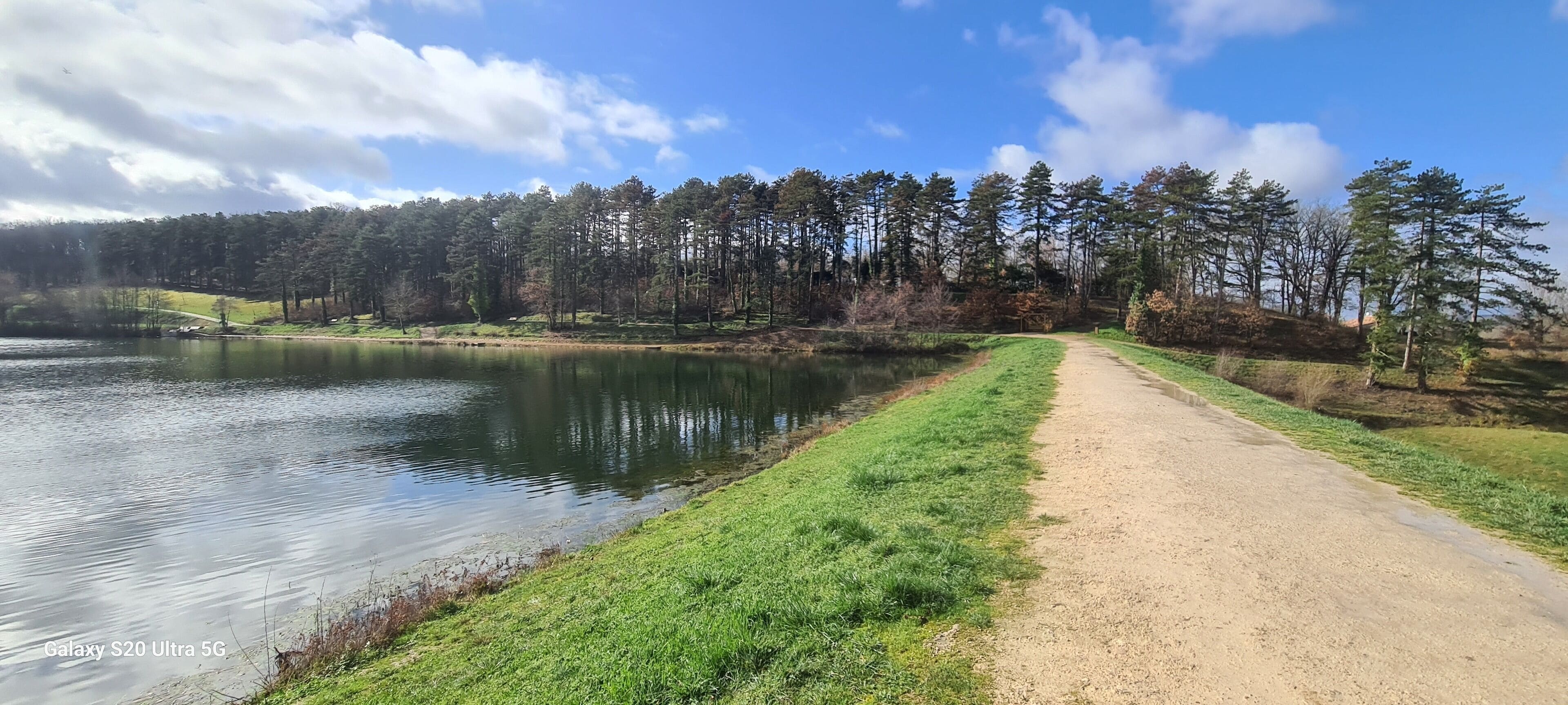 A serene lakeside path with a dirt trail and dense forest in the background.