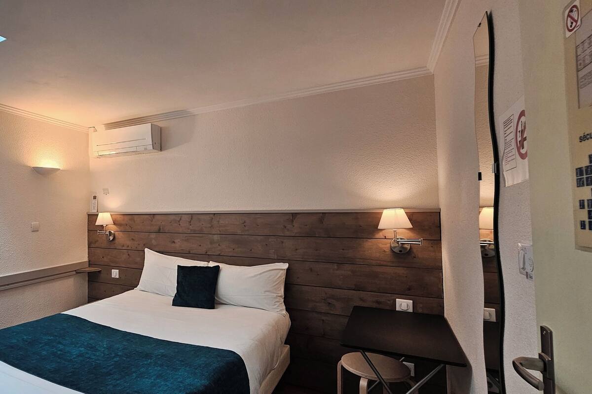 Une chambre d’hôtel avec un lit, une tête de lit en bois, une lucarne et un climatiseur fixé au mur.