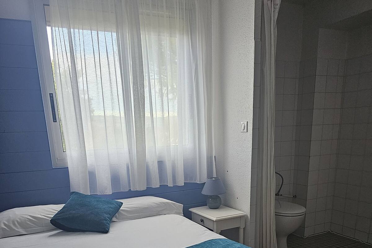 Une chambre à coucher comprenant un lit, une table de chevet, une lampe, une fenêtre avec des rideaux et une vue sur une salle de bain carrelée.