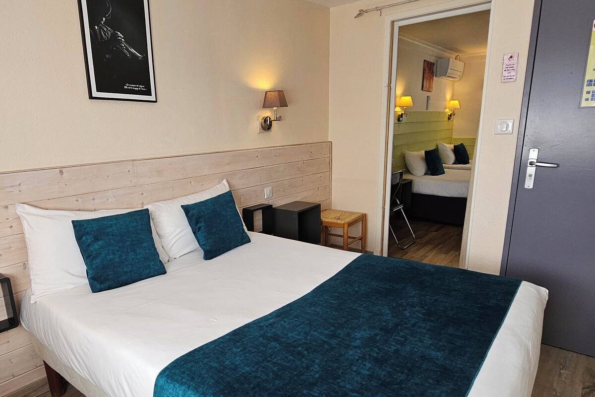 Une chambre d’hôtel avec une tête de lit en bois, un lit avec une literie blanche et turquoise, deux lampes fixées au mur et un tableau encadré accroché au mur.