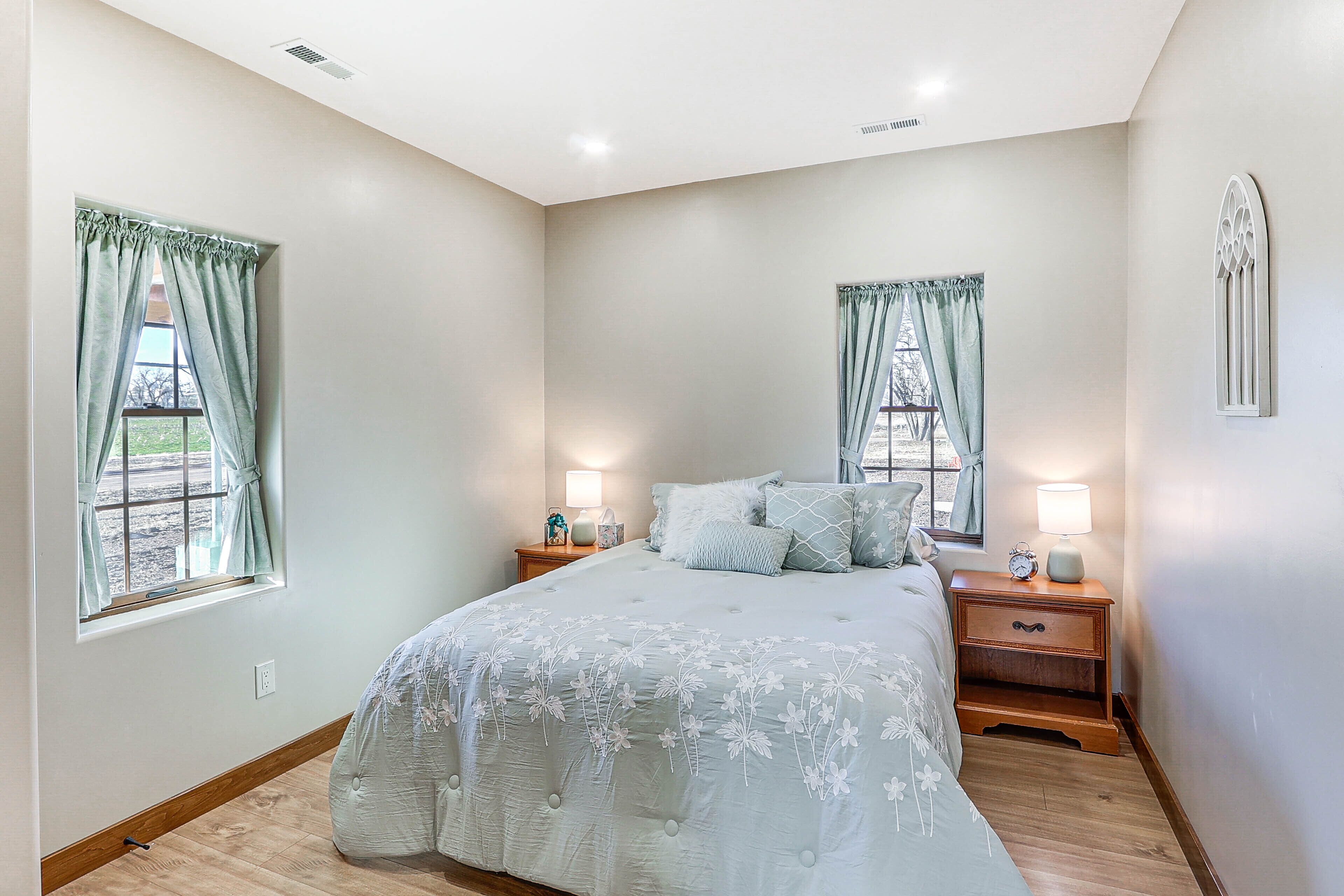 Un dormitorio con una cama grande, dos mesitas de noche, dos ventanas con cortinas, una mesita de noche con lámpara, y un aplique de pared.