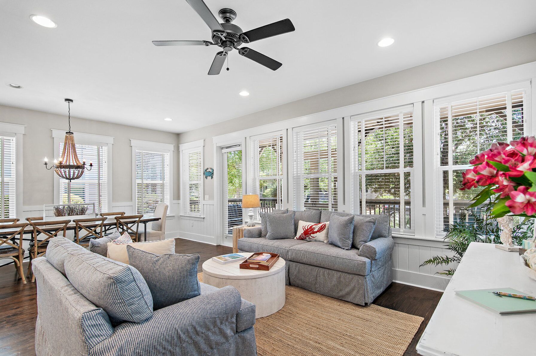 A spacious living room with a ceiling fan, large windows with blinds, a chandelier, and a dining area with a table and chairs.