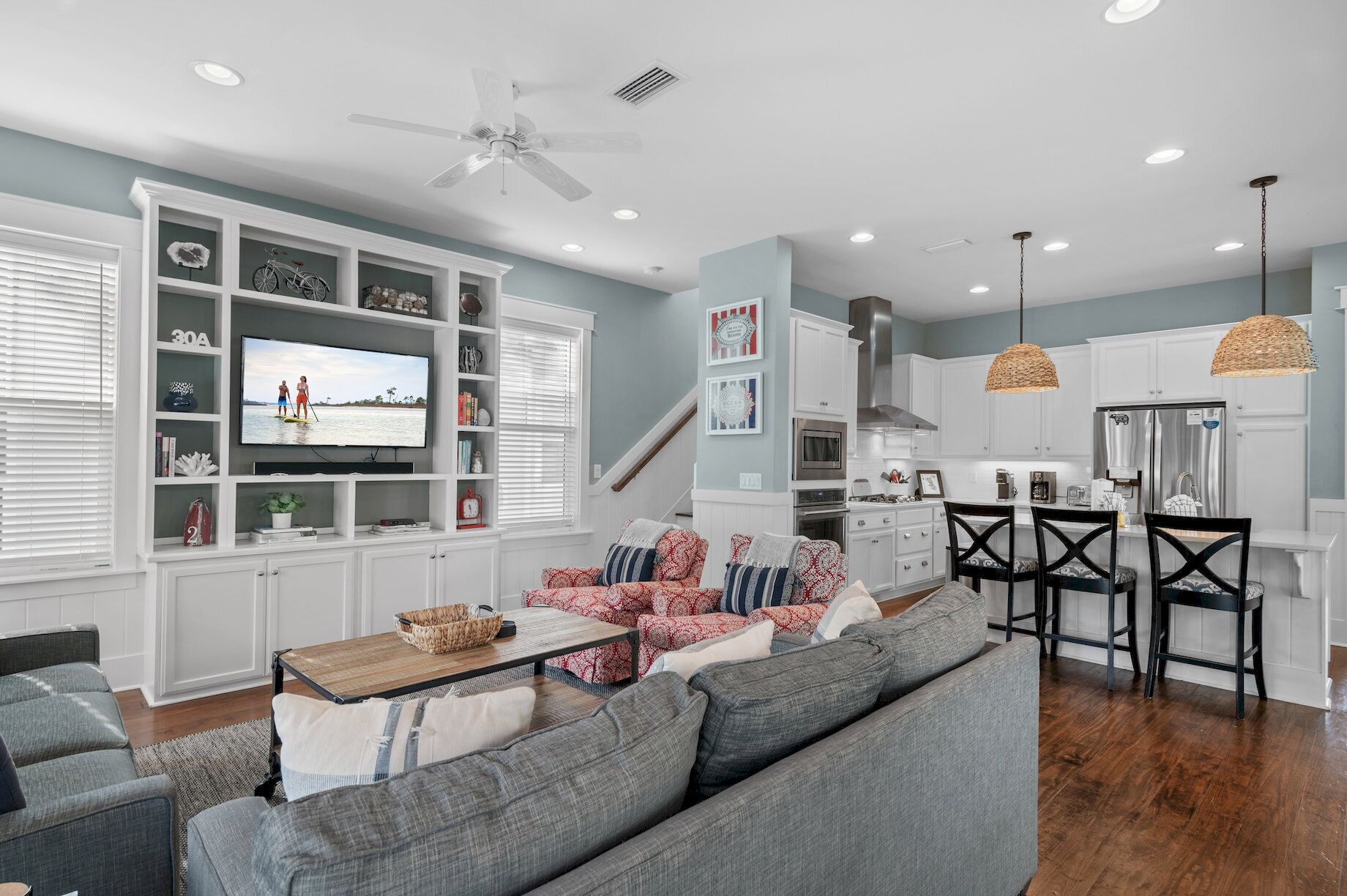 A modern living room with a large TV, grey sofas, a dining area with black chairs, and a kitchen with white cabinets.