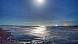 A beach scene with a bright moon and distant city lights. - Billiards & Beaches by Avantstay Cart Boardwalk + Pool Table (Port Aransas)