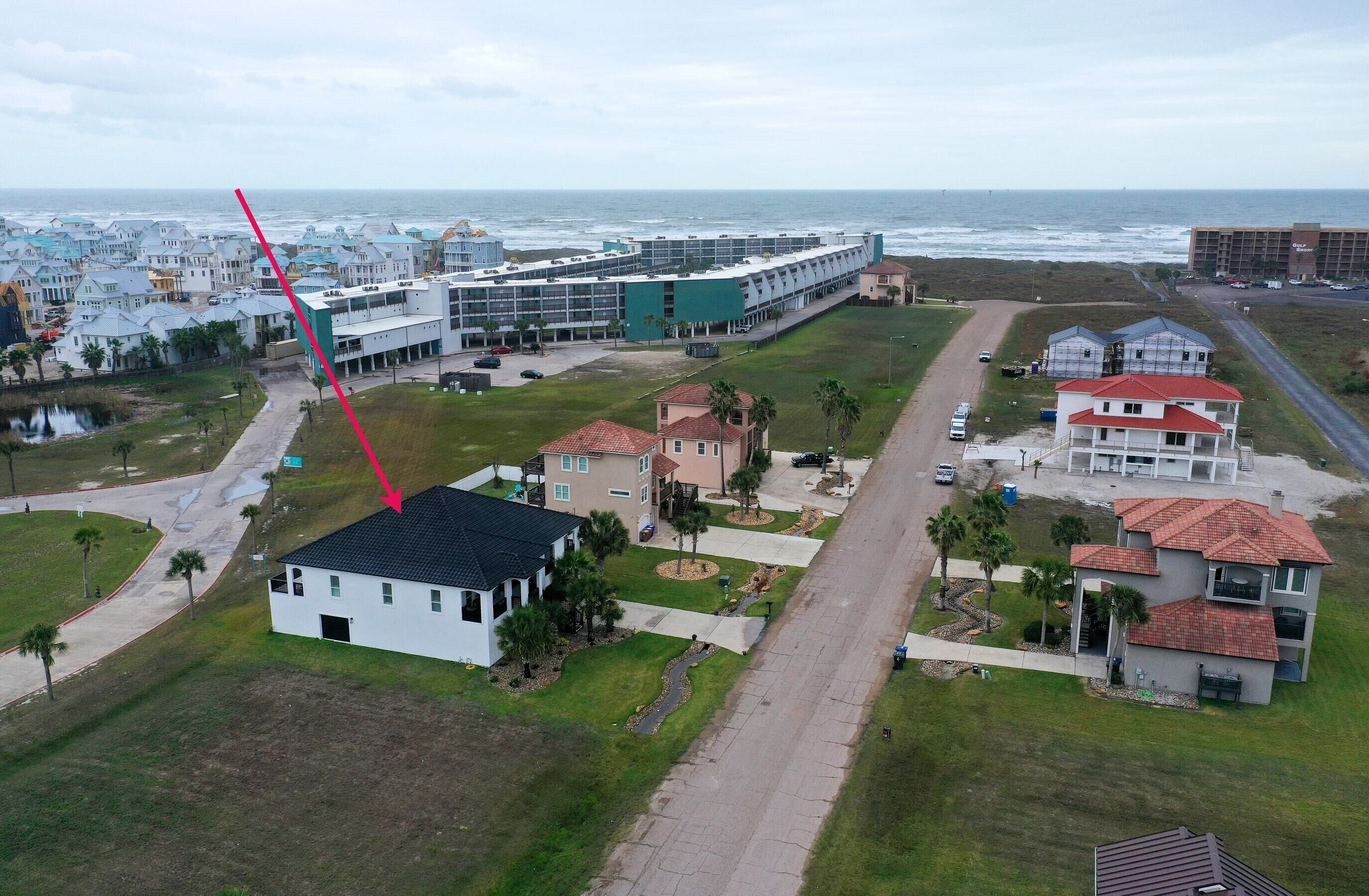 An aerial view of a coastal residential area with a mix of single and multi-story houses, a road, and a beach in the background.