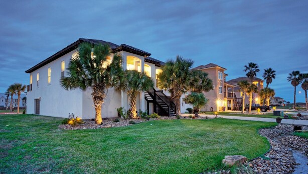 A two-story house with a well-maintained lawn, palm trees, and a staircase leading to the entrance. - Billiards & Beaches by Avantstay Cart Boardwalk + Pool Table (Port Aransas)
