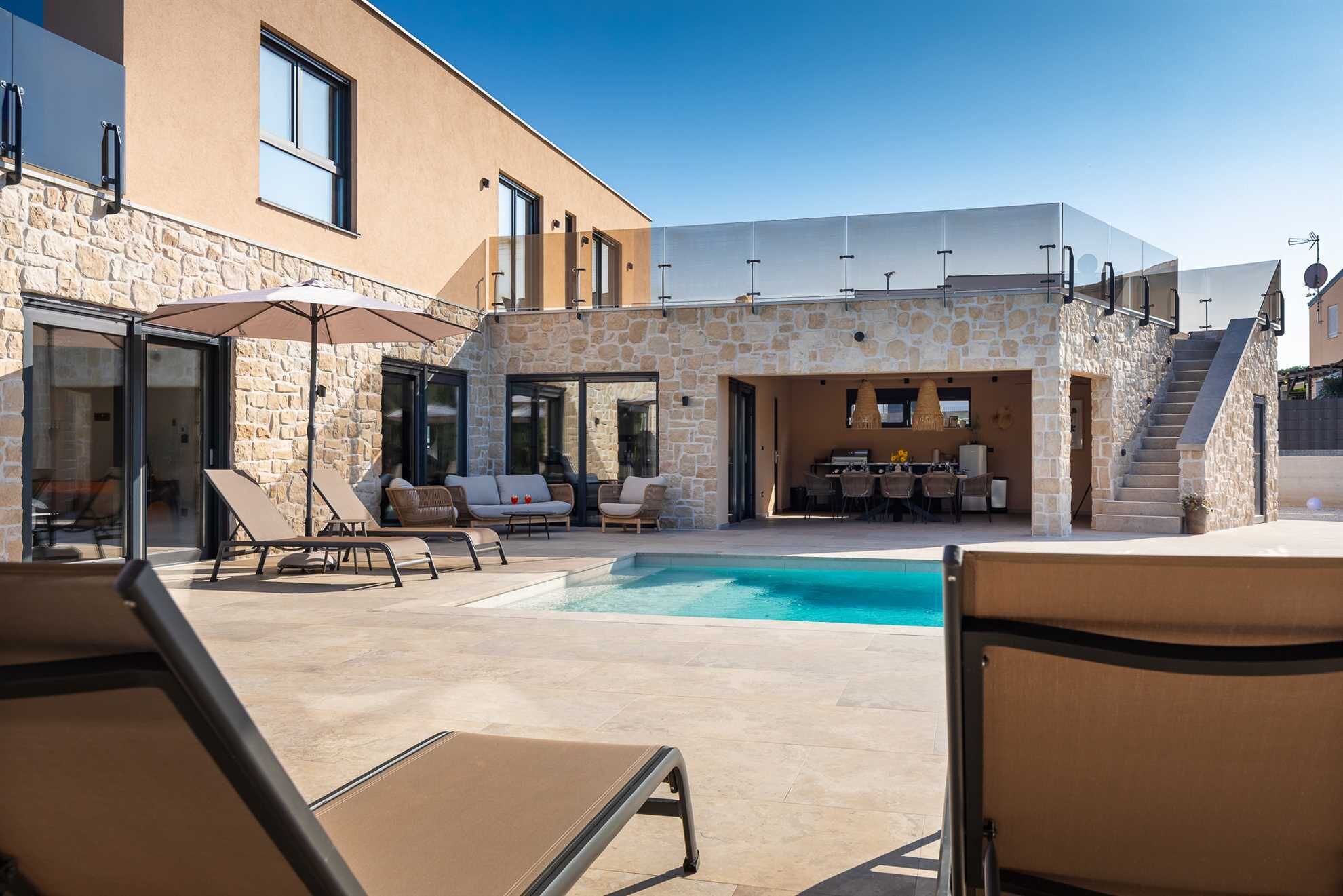 A poolside area with sun loungers, a barbecue setup, and a stone building with a modern design.