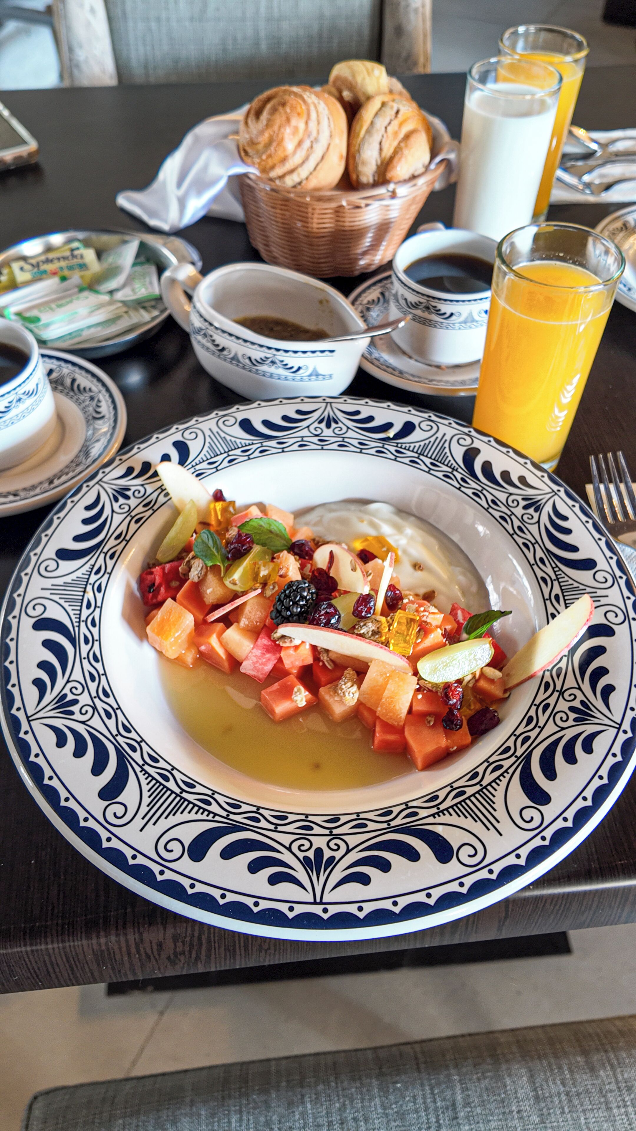 A breakfast setting with a plate of fruit salad, a bowl of soup, and a glass of orange juice on a table.