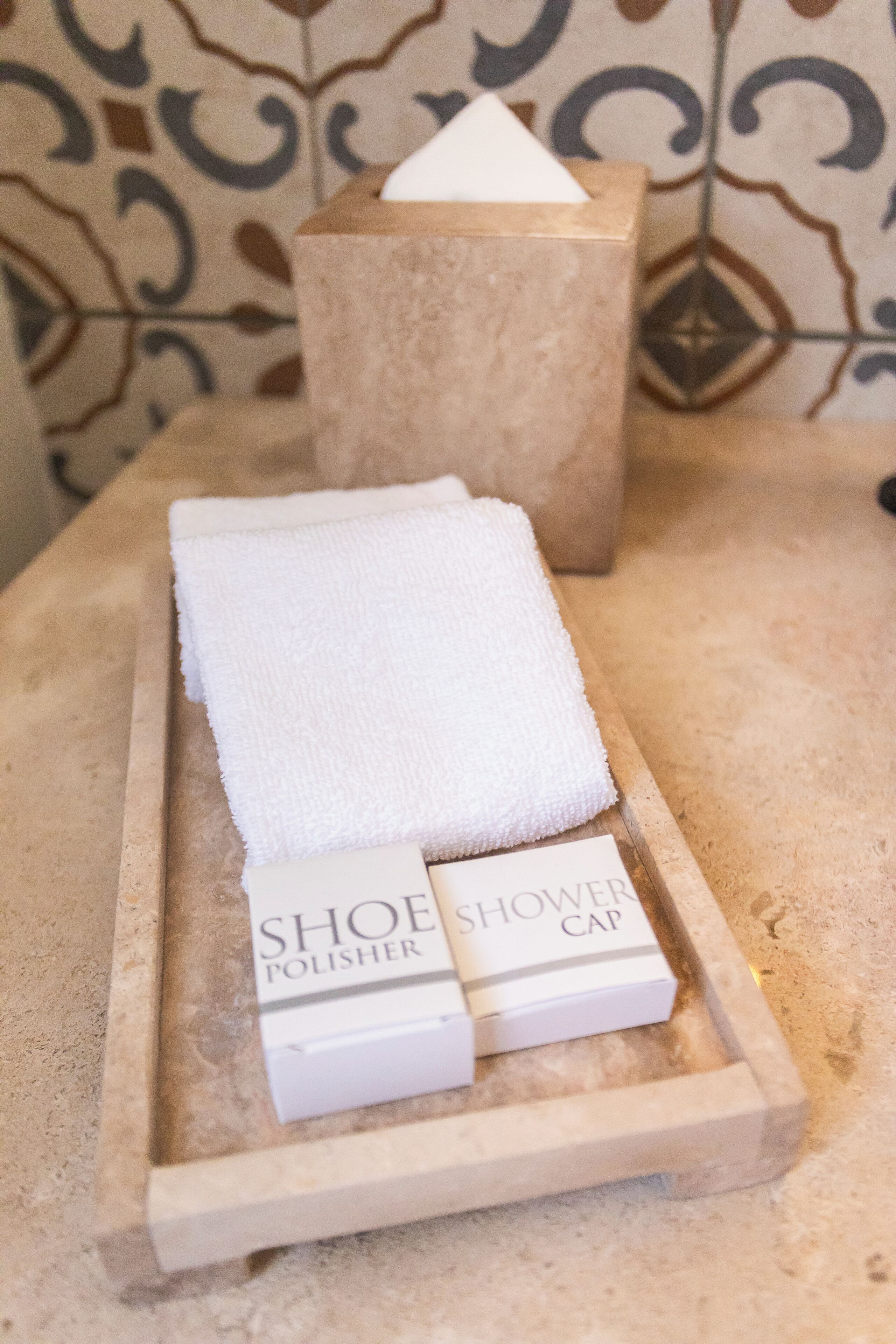 Un plateau avec une serviette blanche et deux boîtes étiquetées « PRODUIT À CIRER LES CHAUSSURES » et « CHARLOTTE DE DOUCHE », posé sur un comptoir à surface texturée.