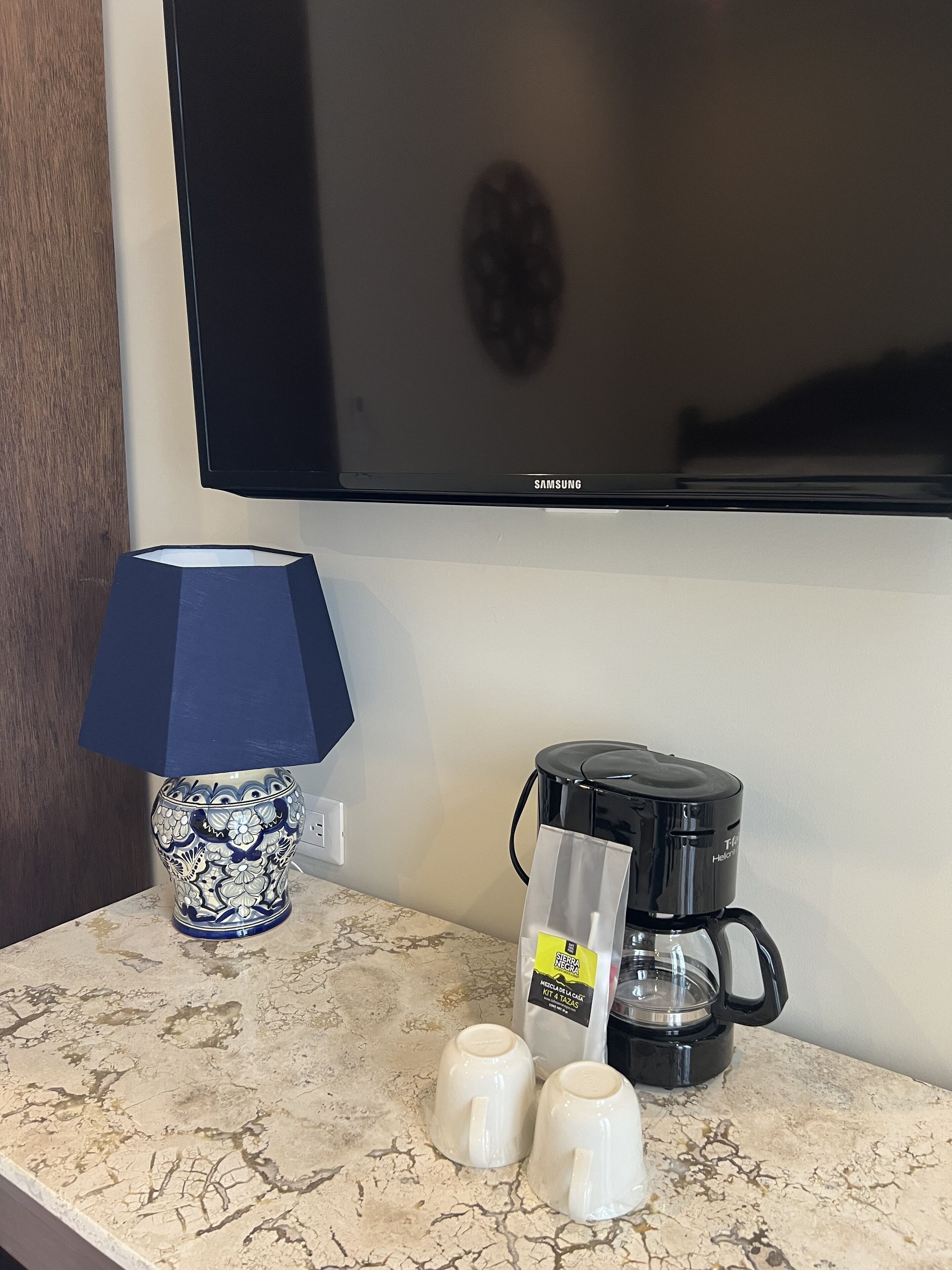 Ein Hotelzimmer mit Flachbildfernseher, Kaffeemaschine, einer blauen und weißen Vase sowie zwei weißen Tassen auf einer Marmorarbeitsplatte.