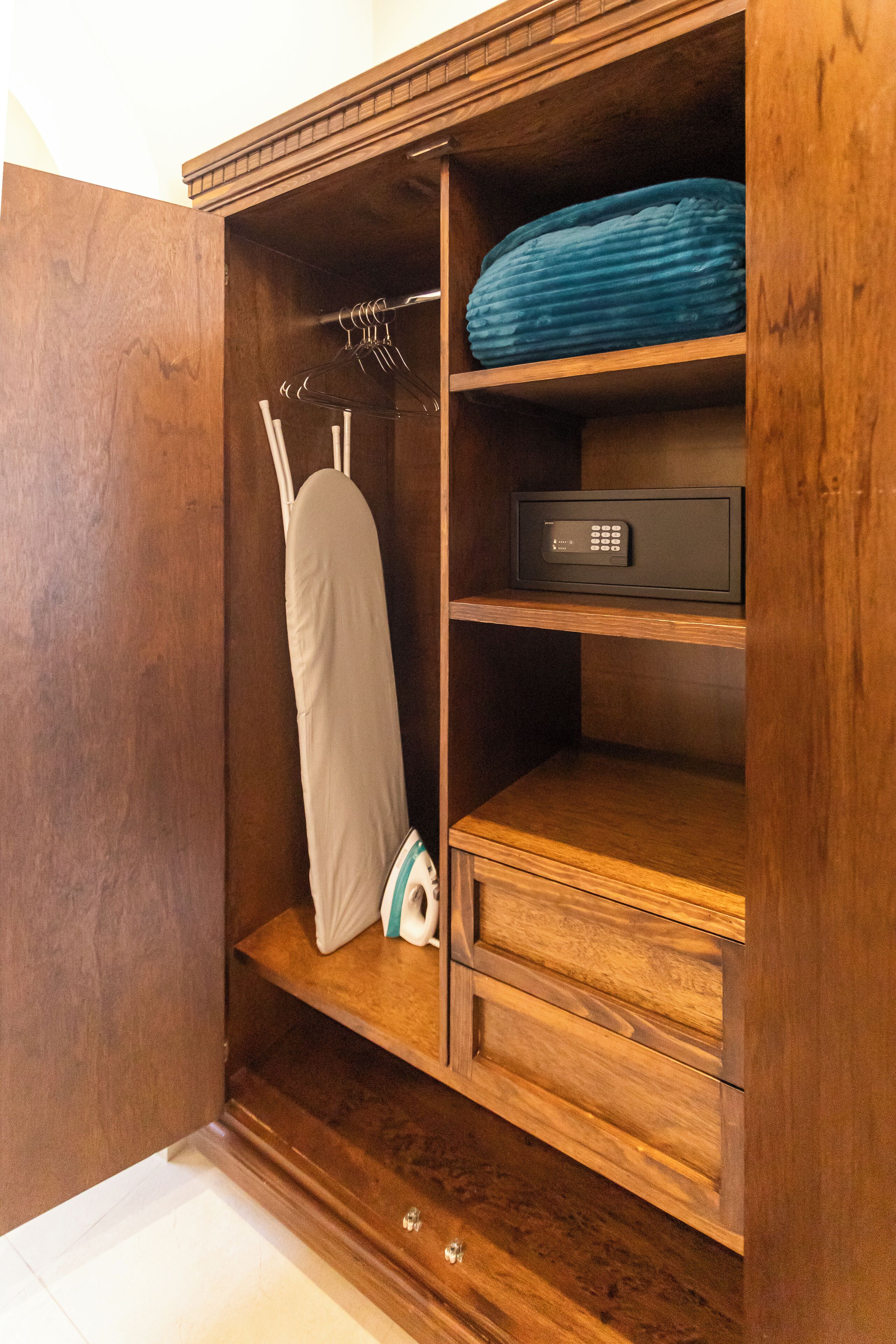 A wooden wardrobe with a hanging iron, a folded blue blanket, and a black electronic device on the top shelf.