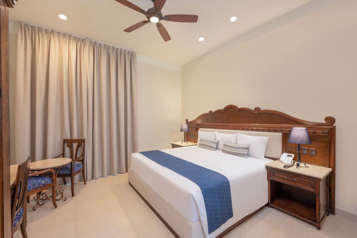 A hotel room with a large bed, a wooden headboard, a bedside table with a lamp, a chair, and a window with curtains.