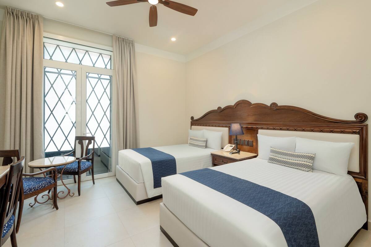 A hotel room with two beds, a dining area with a table and chairs, a ceiling fan, and a window with curtains.