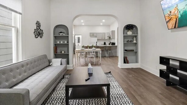 Un salon moderne avec un canapé gris, une table basse et un téléviseur fixé au mur. Au fond, on aperçoit une cuisine avec des meubles blancs et un coin repas.