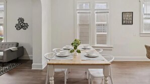 Un coin repas avec une table en bois dressée pour six personnes, des chaises blanches et un vase contenant des feuilles vertes.