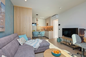 A modern living room with a grey sofa, a TV, and a dining area with chairs and a kitchen in the background. - Hedera Estate, Hedera A44 (Dubrovnik)