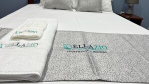 Un lit bien fait, avec une couette blanche et des oreillers gris, une serviette blanche pliée et une couverture grise sur laquelle est imprimé le texte « BELLAZIO APARTMENTS & HOUSING ».