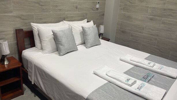 Un lit bien fait, avec une literie blanche et des oreillers gris, une table de chevet avec une lampe et une serviette sur laquelle est inscrit le nom de l’hôtel.