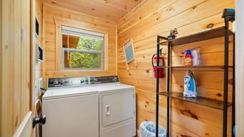 A compact laundry room with a washing machine, a dryer, a shelf with cleaning supplies, and a window with a view of trees.