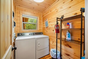 Pequeña lavandería con lavadora, secadora, estante con productos de limpieza y ventana con vista a árboles.