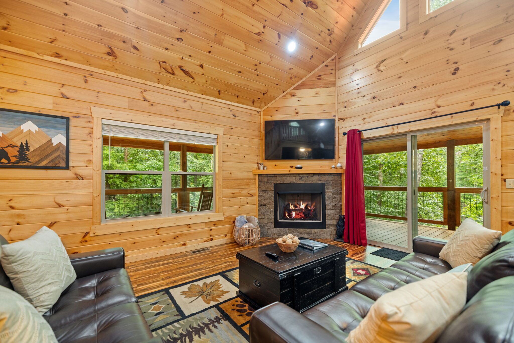 L’intérieur chaleureux d’un chalet avec une cheminée, des canapés en cuir, un téléviseur et de grandes fenêtres offrant une vue sur la forêt.