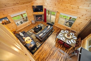 A wooden cabin interior with a living area, fireplace, dining table, and large windows.
